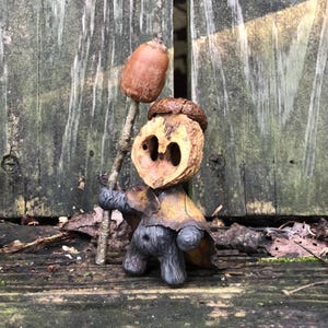 Op de afbeelding: Een grillige sculptuur gemaakt van natuurlijke materialen. De figuur heeft een walnootschaalkop, een kleine eikelhoed en een lichaam gemaakt van een notendop. Het houdt een takje met een eikel vast, wat een uniek, handgemaakt ontwerp laat zien.