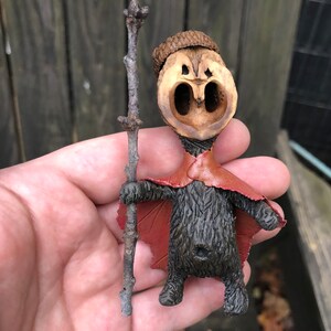 May include: A whimsical, handmade figurine crafted from a walnut shell, featuring a carved face and a small acorn cap. The figure wears a red cape and holds a twig, showcasing a unique, rustic aesthetic. The figurine is held in a hand.
