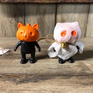 May include: Two Halloween-themed figurines. One is a black figure with an orange pumpkin head. The other is a white ghost cat figure with orange eyes and a small gold bell. Both figures are on a wooden surface.