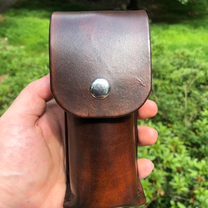 May include: A brown leather case with a silver snap closure. The case is designed to hold a small item, such as a flashlight or a pen.