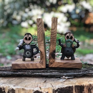 Puede incluir: Un par de sujetalibros de madera con figuras talladas a mano con caras caprichosas. Los sujetalibros están pintados en tonos marrones, negros y verdes. Las figuras sostienen objetos, lo que añade a su carácter único.