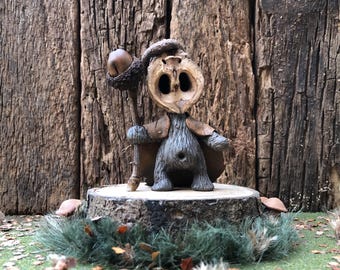 May include: A small, wooden figurine of a creature with a large acorn head, wearing a cloak and holding a staff. The figurine is standing on a wooden stump, surrounded by green grass and fallen leaves.