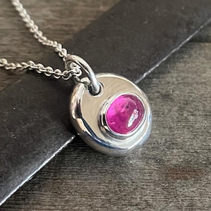 May include: A silver pendant necklace featuring a polished, rounded silver disc with an off-centre, oval-shaped, vibrant pink gemstone set into the metal. The pendant hangs from a delicate silver chain with small, uniform links.