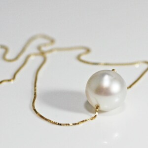 A single round white pearl on a thin gold chain, lying on a white surface.