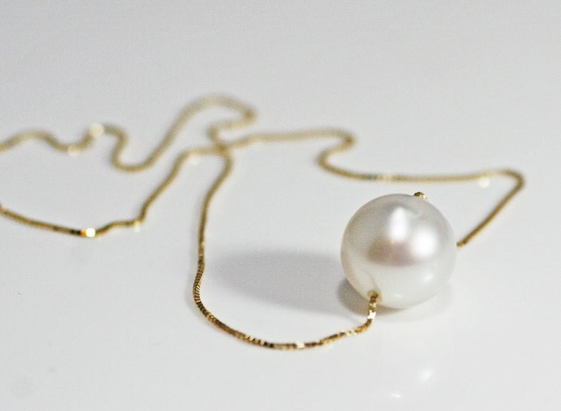 A single round white pearl on a thin gold chain, lying on a white surface.