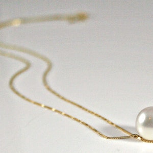 A single round white pearl on a thin gold chain, lying on a white surface.