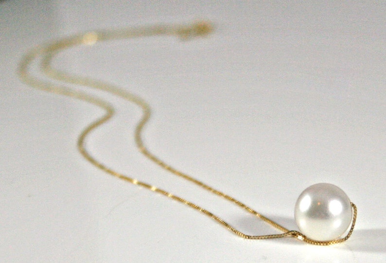 A single round white pearl on a thin gold chain, lying on a white surface.