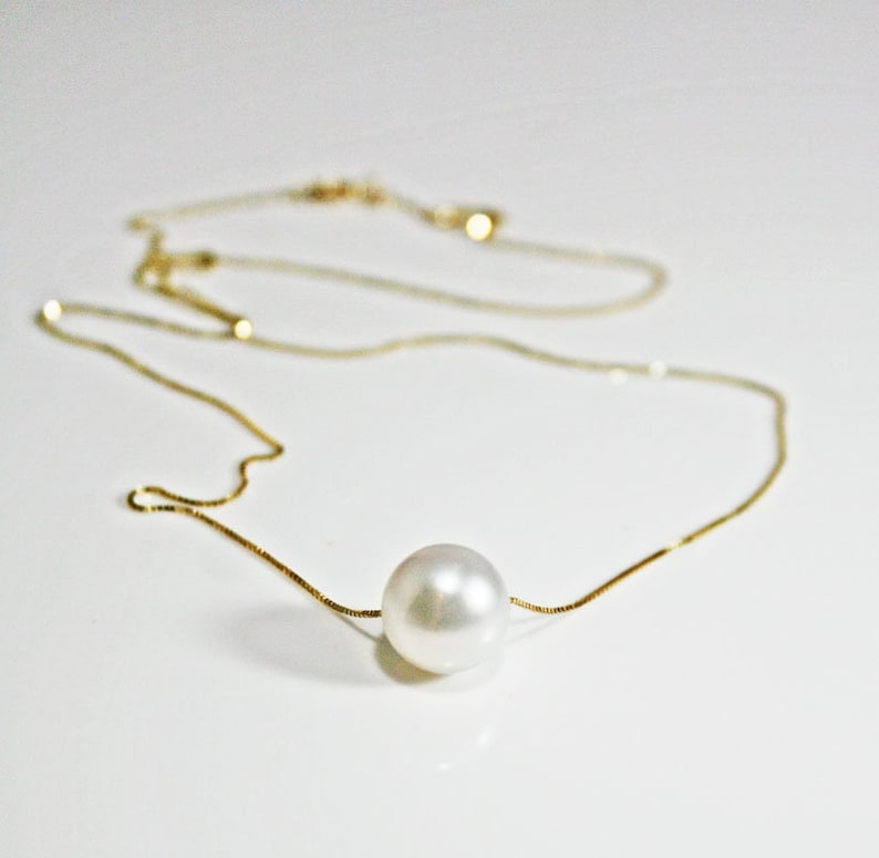 A single round white pearl on a thin gold chain, lying on a white surface.