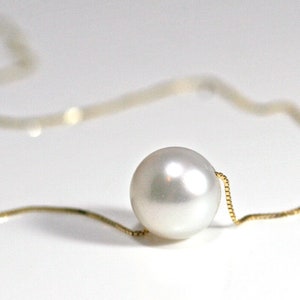 A single round white pearl on a thin gold chain, lying on a white surface.