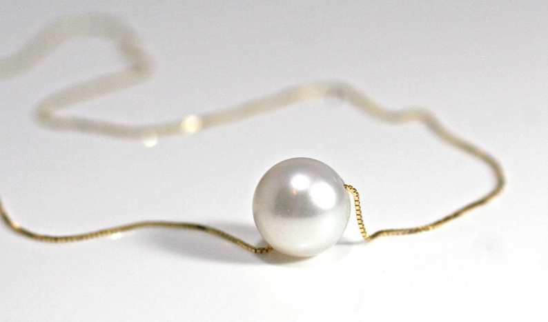 A single round white pearl on a thin gold chain, lying on a white surface.