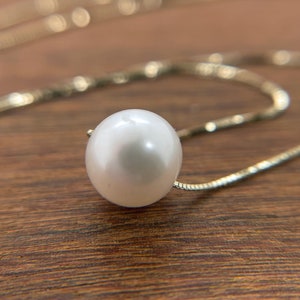 A single round white pearl on a thin gold chain, lying on a wood surface.