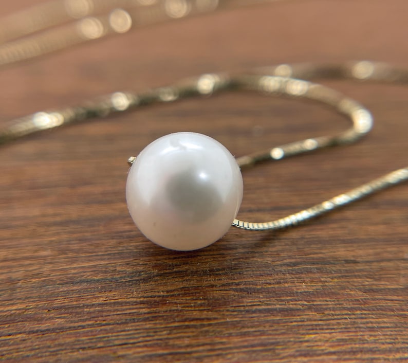 A single round white pearl on a thin gold chain, lying on a wood surface.