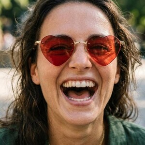 May include: Heart-shaped sunglasses with translucent red lenses and a thin gold-tone metal frame. The eyewear features a classic bridge design connecting the two heart-shaped rims, providing a distinct look with vibrant red tinted lenses.