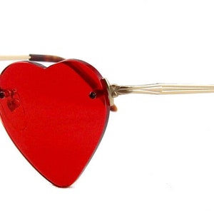 May include: Rimless sunglasses featuring vibrant red heart-shaped lenses. The frames are constructed with thin, metallic gold-toned arms that transition into tortoiseshell-patterned temple tips, providing a distinct contrast to the bold red lenses.