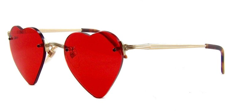 May include: Rimless sunglasses featuring vibrant red heart-shaped lenses. The frames are constructed with thin, metallic gold-toned arms that transition into tortoiseshell-patterned temple tips, providing a distinct contrast to the bold red lenses.