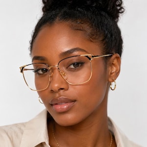 Puede incluir: Gafas de metal en tono dorado con montura cuadrada, bordes de alambre fino y detalles negros en las esquinas superiores. Las gafas cuentan con almohadillas nasales ajustables y patillas delgadas, permitiendo ver claramente los cristales.