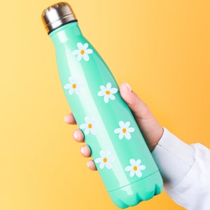 May include: A mint green water bottle with a silver lid, adorned with white daisy flower decals with yellow centers. The bottle is held against a yellow background.