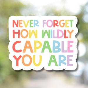 May include: A white sticker with a scalloped edge features the text "NEVER FORGET HOW WILDLY CAPABLE YOU ARE" in rainbow colors. The sticker is designed to be eye-catching and motivational.
