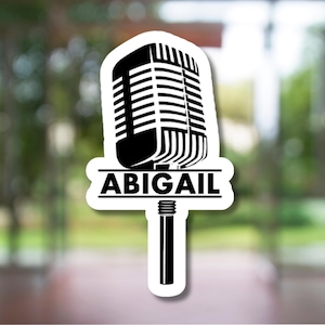 May include: A black and white sticker featuring a vintage microphone design. The name "ABIGAIL" is displayed in bold, black capital letters across the base of the microphone. The sticker has a white border and is set against a blurred background.