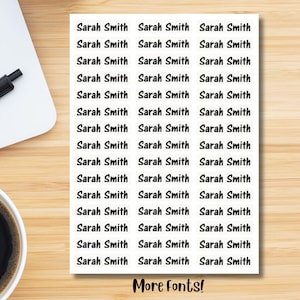 May include: A sheet of white labels printed with the name "Sarah Smith" in a black, simple font. The labels are arranged in rows and columns. The text "More fonts!" is at the bottom of the sheet. The labels are on a light brown wooden surface.