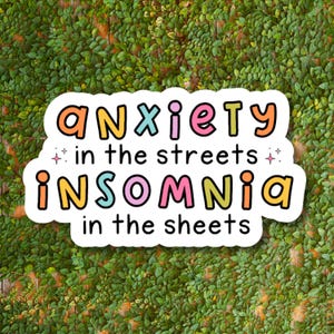 May include: A white sticker with the text "anxiety in the streets insomnia in the sheets" in colorful bubble letters. The sticker is set against a background of green leaves.