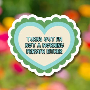May include: A heart-shaped sticker with a scalloped edge. The sticker has a light blue heart inside a green heart, with the text "TURNS OUT I'M NOT A MORNING PERSON EITHER" in black letters. The background is a blurred image of colorful flowers.