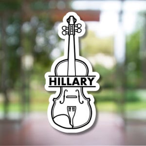 May include: A white sticker in the shape of a violin with the name "HILLARY" in a black, bold font. The sticker has a black outline and is set against a blurred background of green foliage.