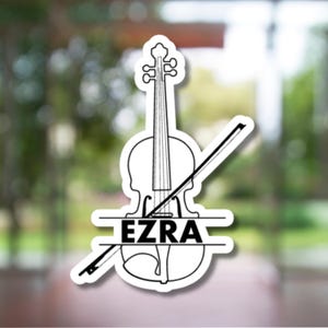 May include: A black and white sticker featuring a violin with a bow. The name "EZRA" is printed in a horizontal bar below the violin. The sticker is set against a blurred green and white background.