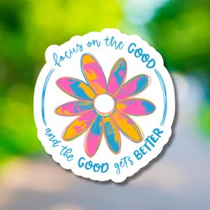 May include: A white sticker with a daisy design. The daisy petals are a mix of pink, yellow, and blue. The text "focus on the GOOD and the GOOD gets BETTER" is written around the flower in blue.