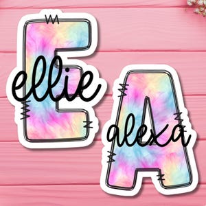 May include: Two colorful tie-dye letters, E and A, with the names "ellie" and "alexa" written in black cursive on them.