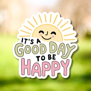 May include: A white sticker with a smiling sun graphic and the text "It's a Good Day to Be Happy" in green and pink. The sun is yellow with orange rays. The sticker has a white border.