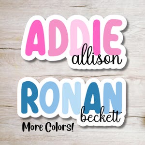May include: Two personalized name stickers with colorful letters. The top sticker is pink and says "ADDIE allison". The bottom sticker is blue and says "RONAN beckett". The text "More Colors!" is below the stickers.
