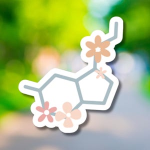 May include: A white sticker featuring a light blue serotonin molecule design, adorned with peach and pink floral accents. The sticker has a white border and is set against a blurred green and white background.