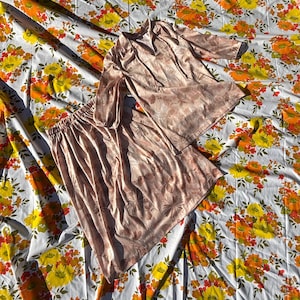 May include: A vintage two-piece outfit in a light pink floral pattern. The set includes a long-sleeved top with a V-neck and a matching skirt. The outfit is displayed on a floral patterned sheet with yellow, orange, and white flowers.