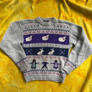 May include: A heather gray long-sleeve sweater featuring a crew neckline and a multi-colored knit pattern. The design includes rows of letters, numbers, white sheep on a purple band, white rabbits on a dark blue band, and three figures.