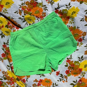 May include: Bright green athletic shorts with an elastic waistband. The shorts are made of a lightweight, possibly nylon, material. The shorts are displayed on a floral patterned fabric with orange, yellow, and red flowers.