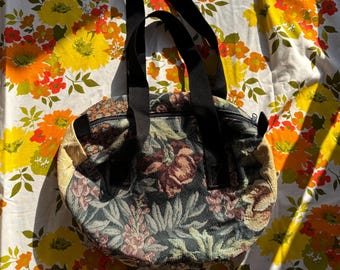 May include: A rounded handbag featuring a dark tapestry fabric with a multi-colored floral pattern, black nylon straps, and a top zipper closure. The sides of the bag are made of a textured, light beige material, providing a contrast to the floral body.