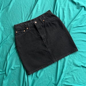 May include: A black denim mini skirt featuring a classic five-pocket design, a button fly closure, and copper-toned metal rivets. The skirt has a straight hemline and visible stitching details along the waistband, pockets, and front panel seams.
