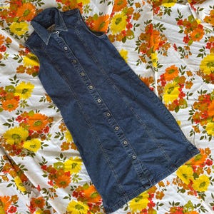 May include: A sleeveless, button-down denim dress in a medium wash. The dress features a collared neckline and a fitted silhouette. It is displayed on a vintage floral patterned fabric with yellow, orange, and white flowers.