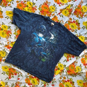May include: A dark blue short-sleeve t-shirt featuring a graphic print of dolphins swimming in an ocean scene. The design includes a bright light reflection on the water surface, multiple dolphins, and underwater coral elements on a mottled fabric.