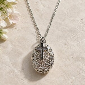 May include: A silver-toned necklace featuring an oval-shaped locket with an intricate, openwork filigree pattern. A small, dark silver-toned cross charm is attached to the top of the locket, which hangs from a delicate silver-toned metal chain.