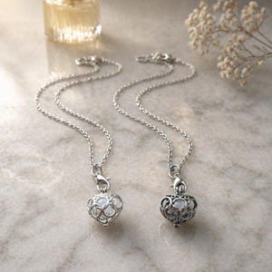 May include: Two silver necklaces with heart-shaped pendants. The pendants have an open filigree design and a light-colored stone inside. The necklaces are displayed on a marble surface, with a small perfume bottle and dried flowers in the background.