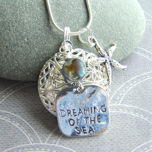 May include: Silver necklace with a silver charm that says "Dreaming of the Sea". The charm is attached to a silver starfish and a silver cage with a blue and green abalone shell.