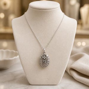 May include: An oval silver locket pendant suspended on a delicate silver chain. The locket features an intricate filigree pattern with a raised, dimensional flower design centered on the front surface, showcasing a polished metallic finish.