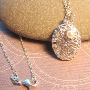 May include: A silver filigree pendant necklace with a small white pearl accent. The pendant is oval shaped and has a delicate, intricate design. The chain is silver and has a lobster clasp.