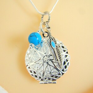 May include: A silver-toned circular locket pendant featuring an intricate filigree cutout design. The locket is accented with a round, bright blue turquoise-colored bead and a silver-toned feather charm with a small blue inlay, hanging from a chain.