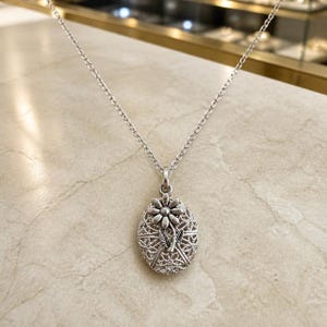 May include: A silver-toned oval locket pendant suspended from a matching delicate link chain. The locket features an intricate, openwork filigree pattern across its surface, topped with a raised, detailed flower motif with leaves.