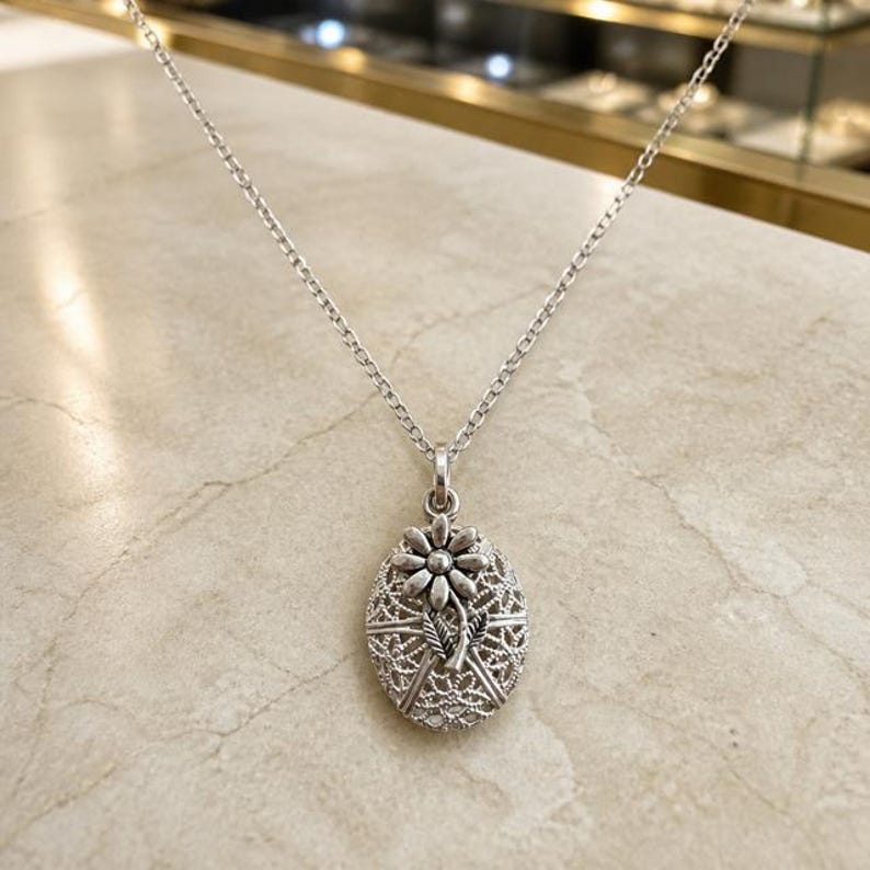 May include: A silver-toned oval locket pendant suspended from a matching delicate link chain. The locket features an intricate, openwork filigree pattern across its surface, topped with a raised, detailed flower motif with leaves.