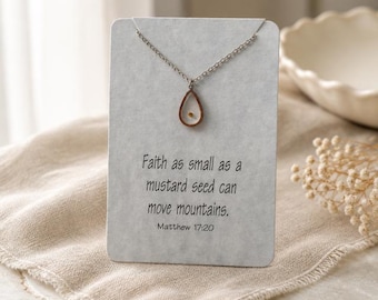 May include: A silver-toned necklace featuring a teardrop-shaped pendant containing a single mustard seed. The necklace is displayed on a rectangular white card printed with the text: Faith as small as a mustard seed can move mountains. Matthew 17:20.