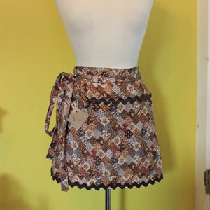 May include: A brown and cream patchwork apron with a scalloped edge and a brown tie. The apron has a pocket on the front.